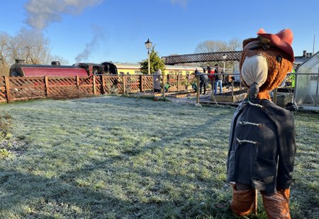 outdoors photo taken on a frosty day. In the foreground is a large teddy bear like a scarecrow. In the background is a steam train