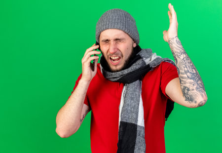 Frustrated young man wearing a red shirt and woolly hat and scarf making a phone call. 