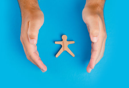 Photograph of two hands cupped in a protective way around a cut-out human figure, all against a plain blue background