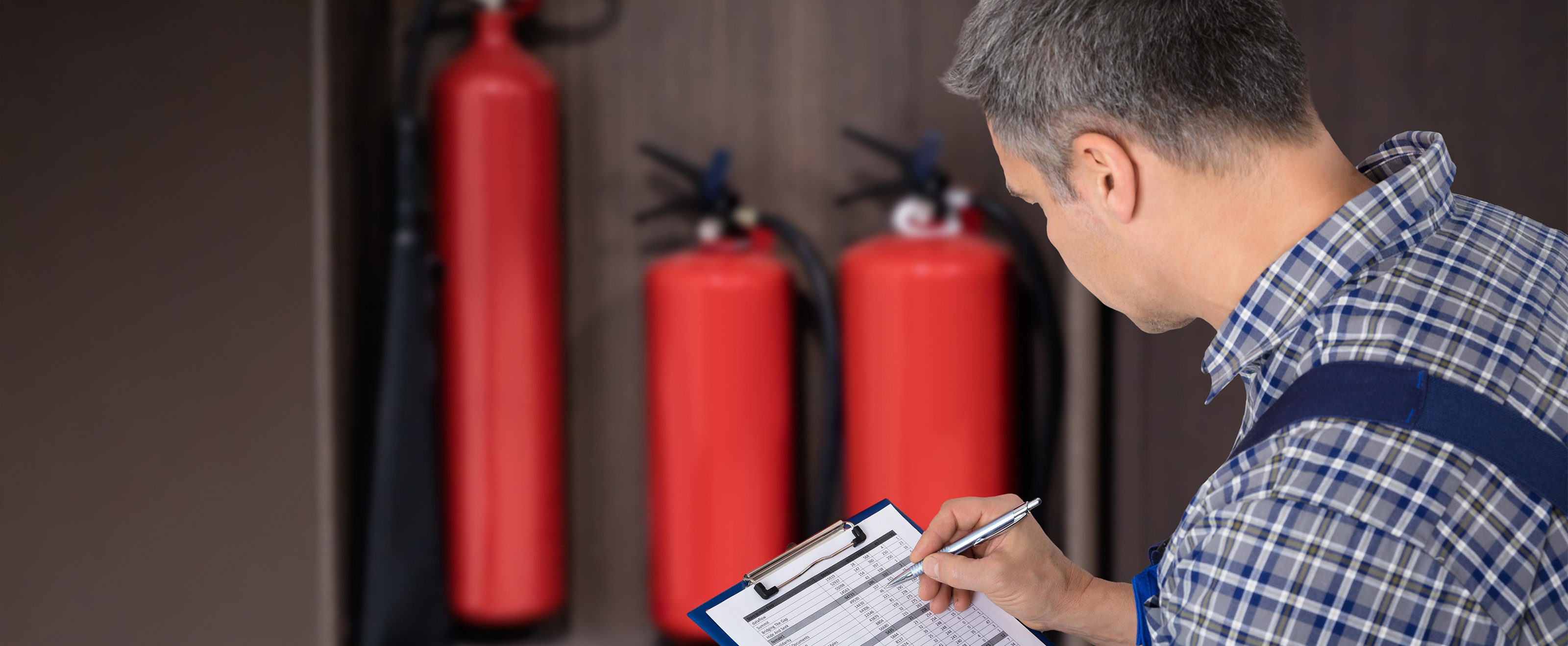 Photo of a man writing notes on a form on a clipboard. In the background we can see three fire extinguishers.