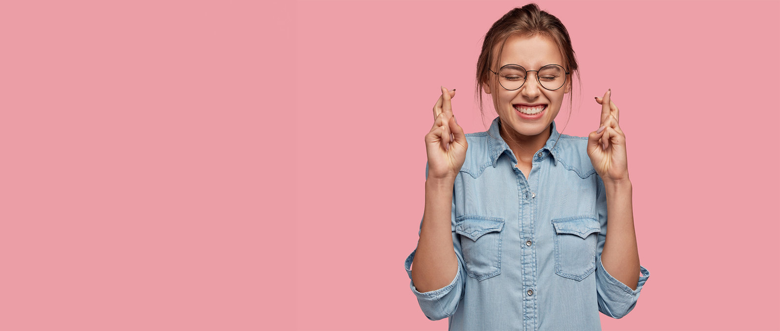 Photo of a young woman with her eyes closed and fingers crossed in front of a pink background