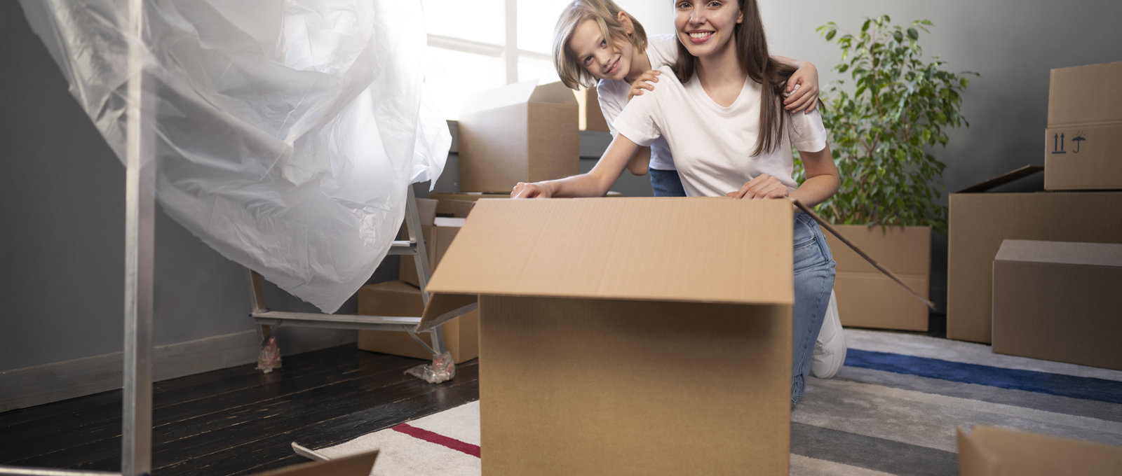 Photo of a young woman and young child smiling at the camera. They are kneeling behind a large cardboard box and surrounded by other boxes