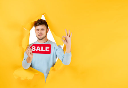 A yellow paper background with a young man bursting through a tear holding up a red sale sign and making an okay sign with his free hand