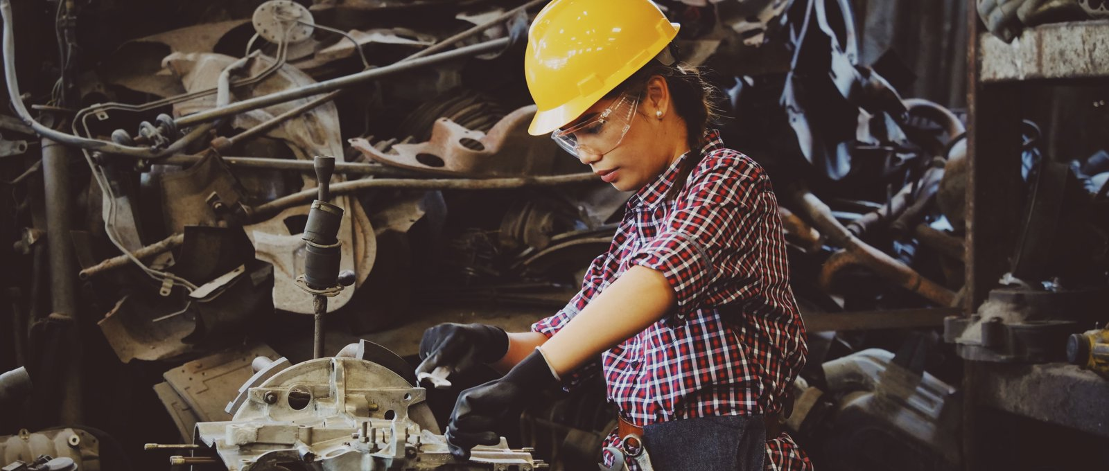 A woman in a check shirt, gloves, a yellow hard hat and safety goggles, fixing a car part using a spanner. She has a navy tool belt at her waist and is surrounded by other car parts.
