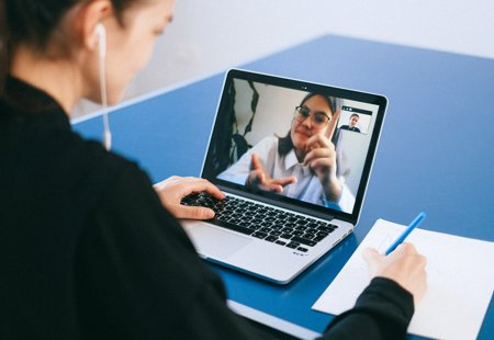 A woman in a black jacket sat at a table in front of a laptop making notes. Another woman in glasses is visible on the laptop screen as part of a video call.