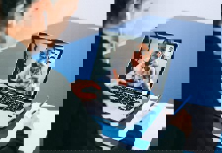 A woman in a black jacket sat at a table in front of a laptop making notes. Another woman in glasses is visible on the laptop screen as part of a video call.