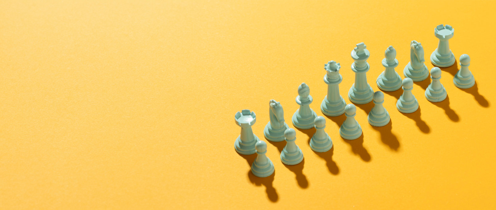 Photograph of a full set of white chess pieces, arranged as they would be for a new game, on a yellow background with strong shadows for each piece