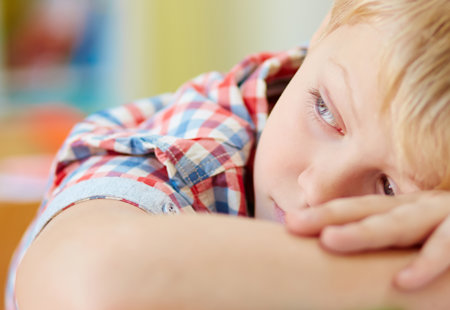 Photograph of a young boy resting his head on his arms on a table and looking sad
