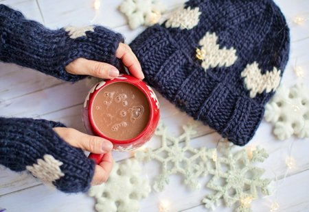 Hands wearing over-the-thumb knitted navy gloves holding a hot chocolate in a large red mug, beside a navy hat and snowflake decorations.