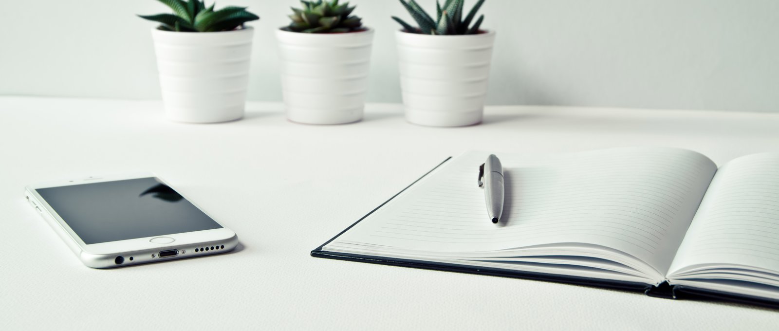 A notebook and pen sit on a white desk beside an iPhone and three small succulents in white pots. 