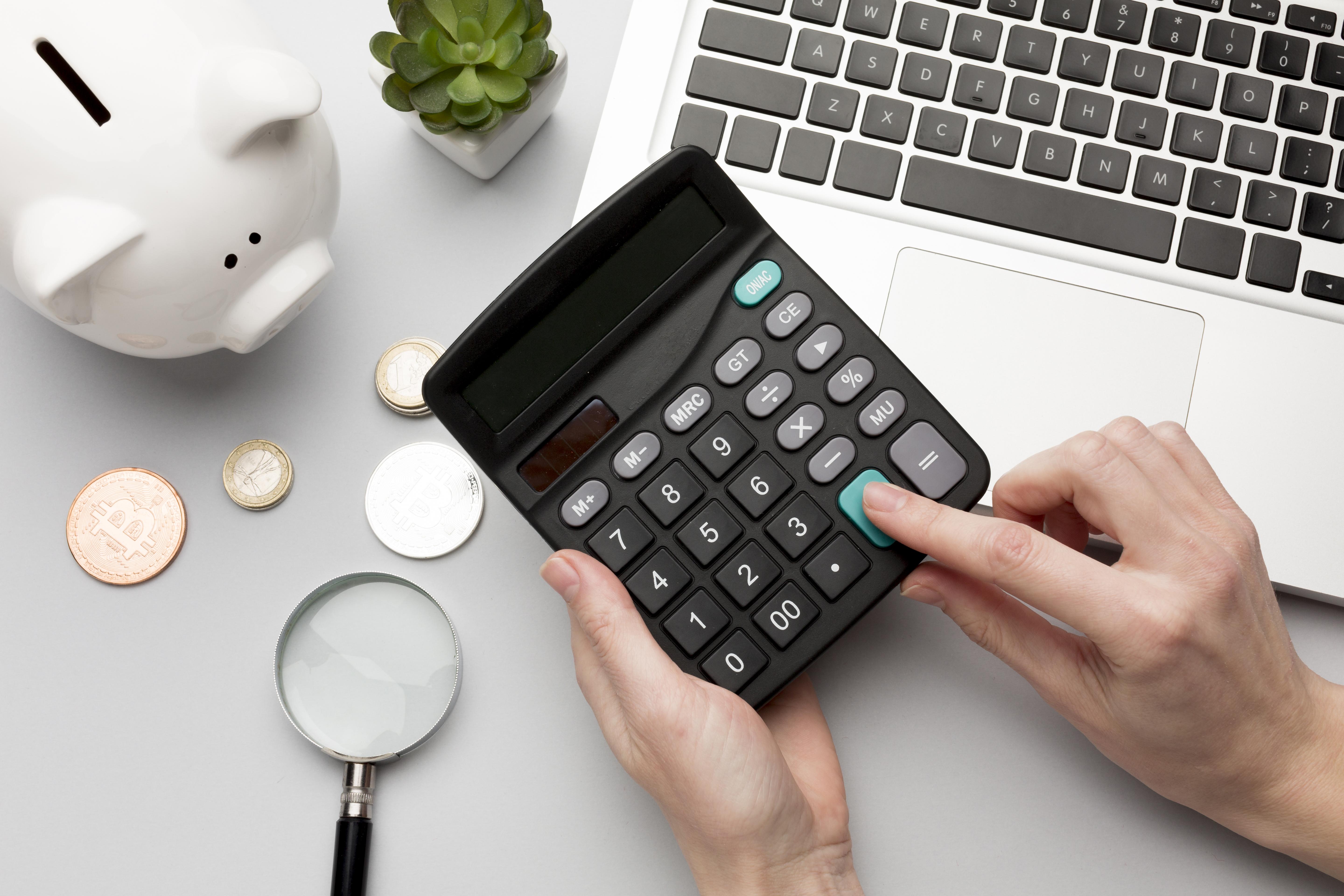 A photo showing hands holding a calculator above a desk, with a white piggy bank, money, a magnifying glass and laptop in the background.