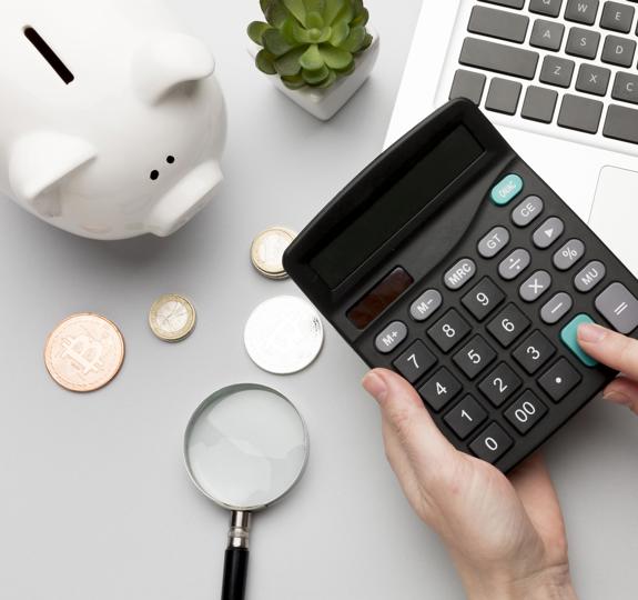 A photo showing hands holding a calculator above a desk, with a white piggy bank, money, a magnifying glass and laptop in the background.