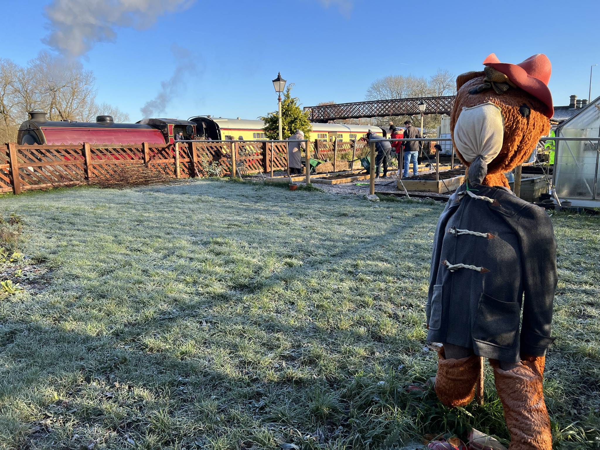 outdoors photo taken on a frosty day. In the foreground is a large teddy bear like a scarecrow. In the background is a steam train