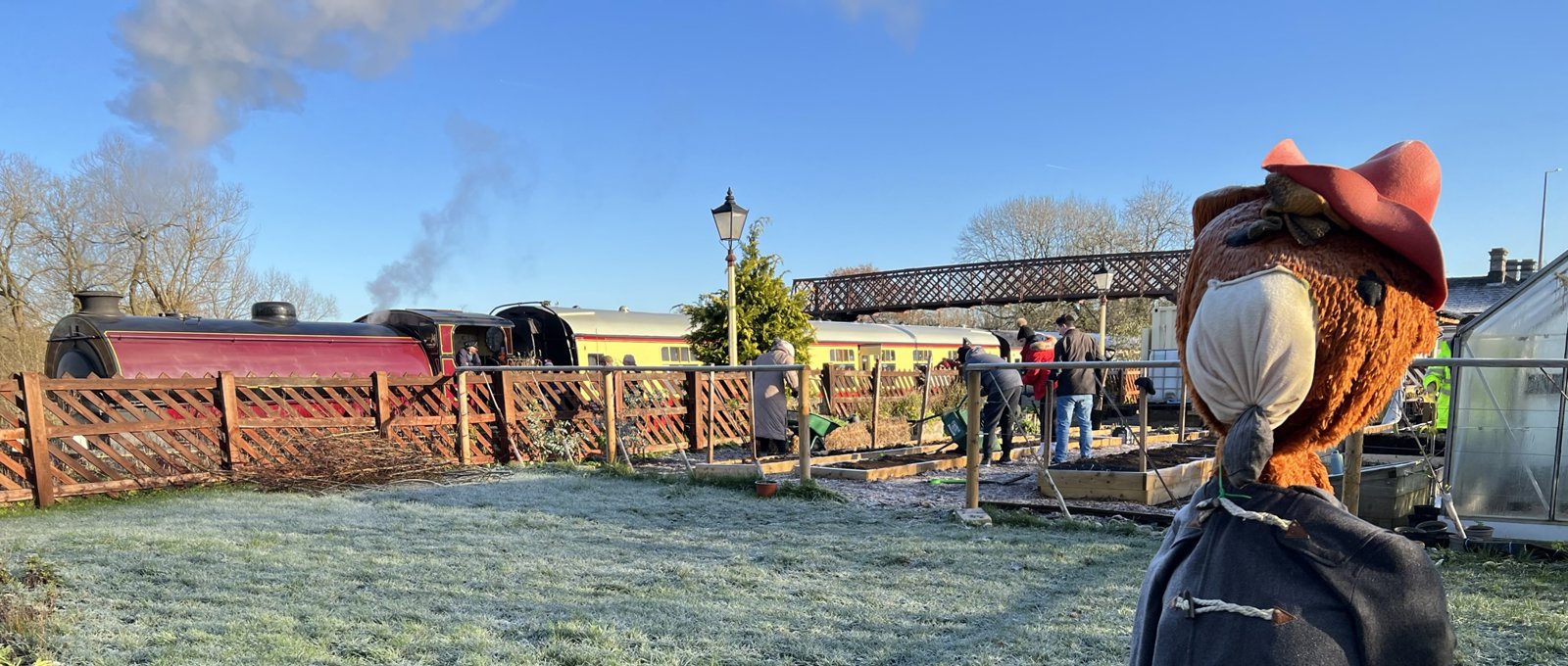 outdoors photo taken on a frosty day. In the foreground is a large teddy bear like a scarecrow. In the background is a steam train