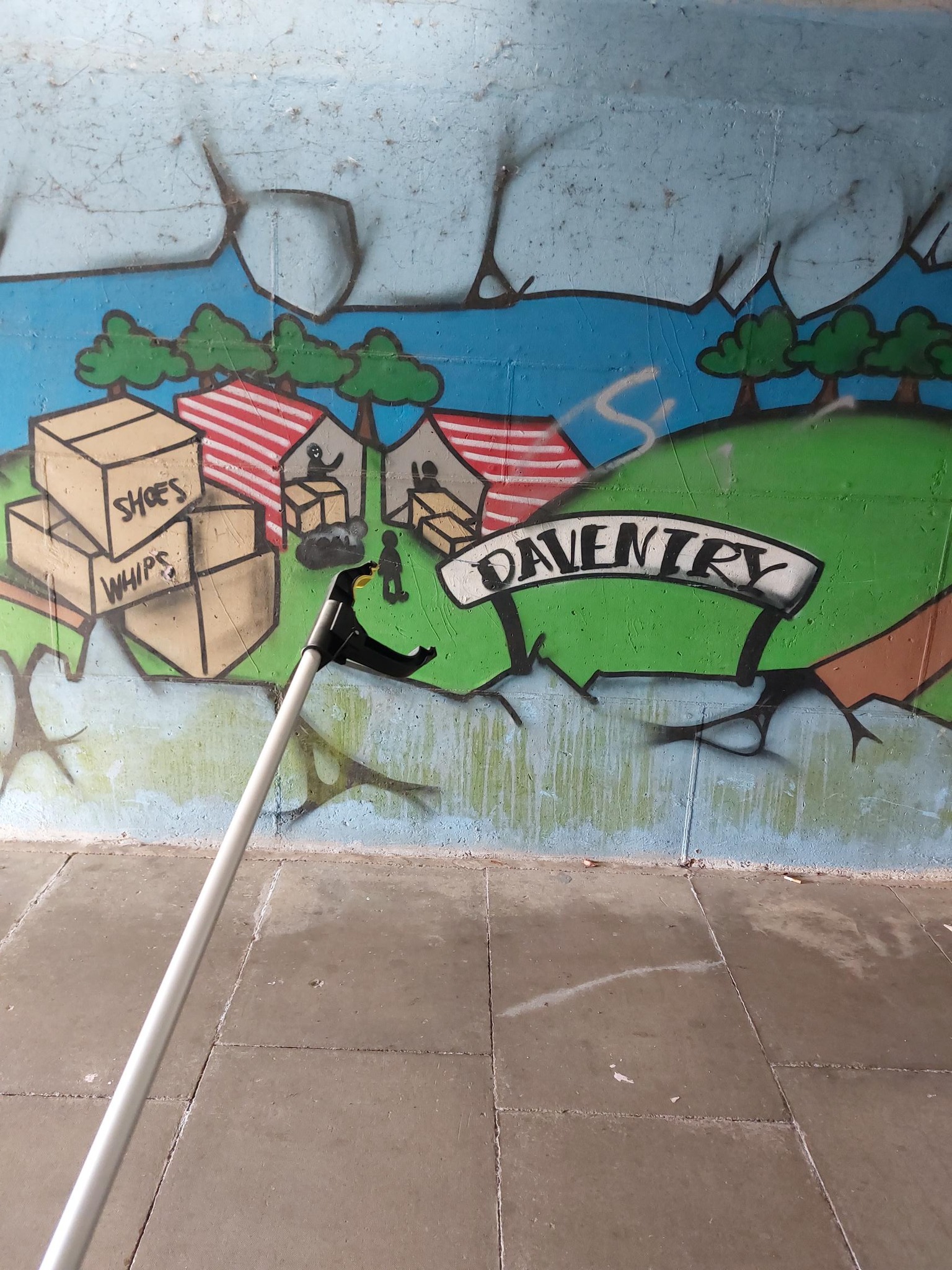 A photo of a litter picker tool in front of graffiti on a wall. The graffiti has a sign saying 'Daventry' with fields and people in tents and cardboard boxes.