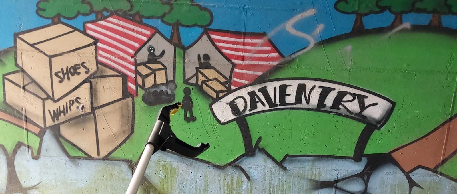 A photo of a litter picker tool in front of graffiti on a wall. The graffiti has a sign saying 'Daventry' with fields and people in tents and cardboard boxes.