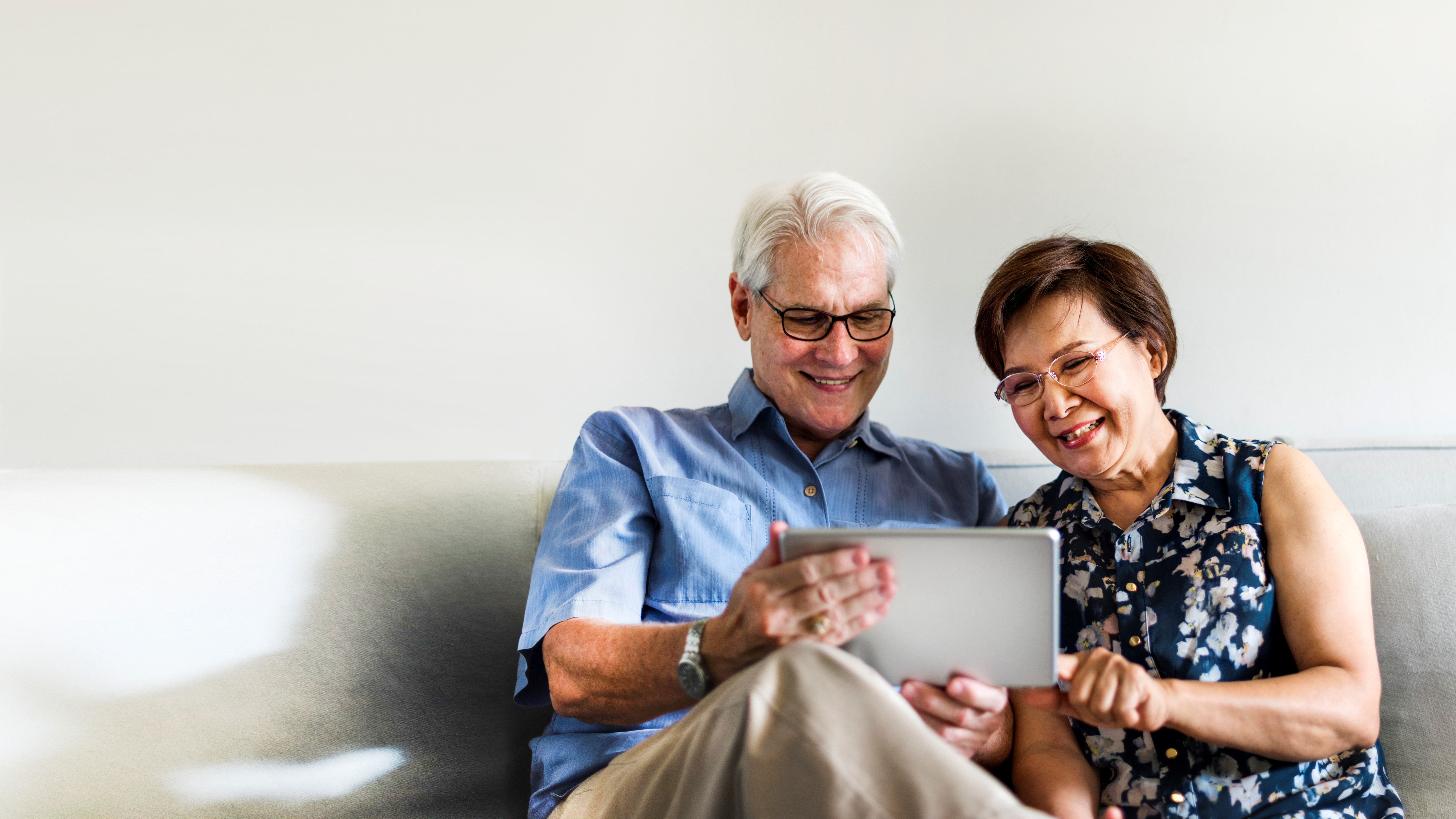 Photo of an older couple using a tablet in their living room. Man is to the left, he's got white hair, glasses and wearing a blue shirt with cream trousers. Women is to the right, she has short brown hair, glasses and wearing a blue, flowery shirt. The man is holding the silver tablet while the woman is pointing at it, while they're sat on a cream sofa with a white/cream wall behind them.