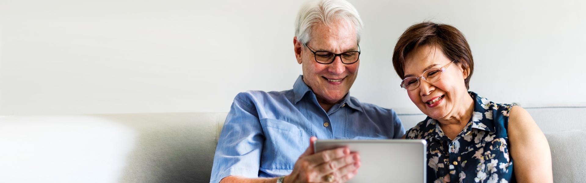 Photo of an older couple using a tablet in their living room. Man is to the left, he's got white hair, glasses and wearing a blue shirt with cream trousers. Women is to the right, she has short brown hair, glasses and wearing a blue, flowery shirt. The man is holding the silver tablet while the woman is pointing at it, while they're sat on a cream sofa with a white/cream wall behind them.