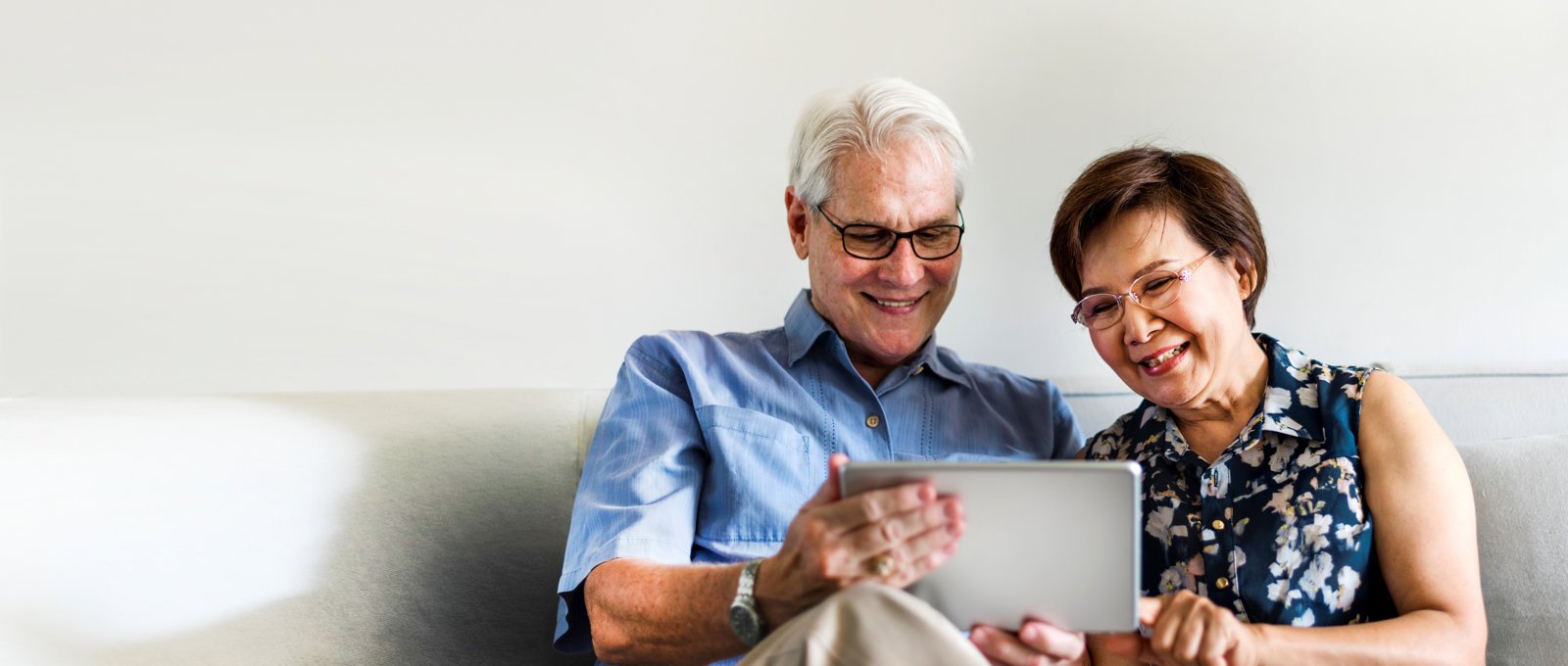 Photo of an older couple using a tablet in their living room. Man is to the left, he's got white hair, glasses and wearing a blue shirt with cream trousers. Women is to the right, she has short brown hair, glasses and wearing a blue, flowery shirt. The man is holding the silver tablet while the woman is pointing at it, while they're sat on a cream sofa with a white/cream wall behind them.