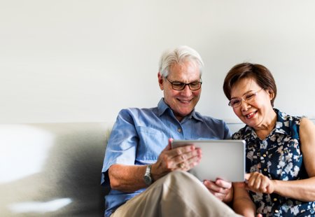 Photo of an older couple using a tablet in their living room. Man is to the left, he's got white hair, glasses and wearing a blue shirt with cream trousers. Women is to the right, she has short brown hair, glasses and wearing a blue, flowery shirt. The man is holding the silver tablet while the woman is pointing at it, while they're sat on a cream sofa with a white/cream wall behind them.