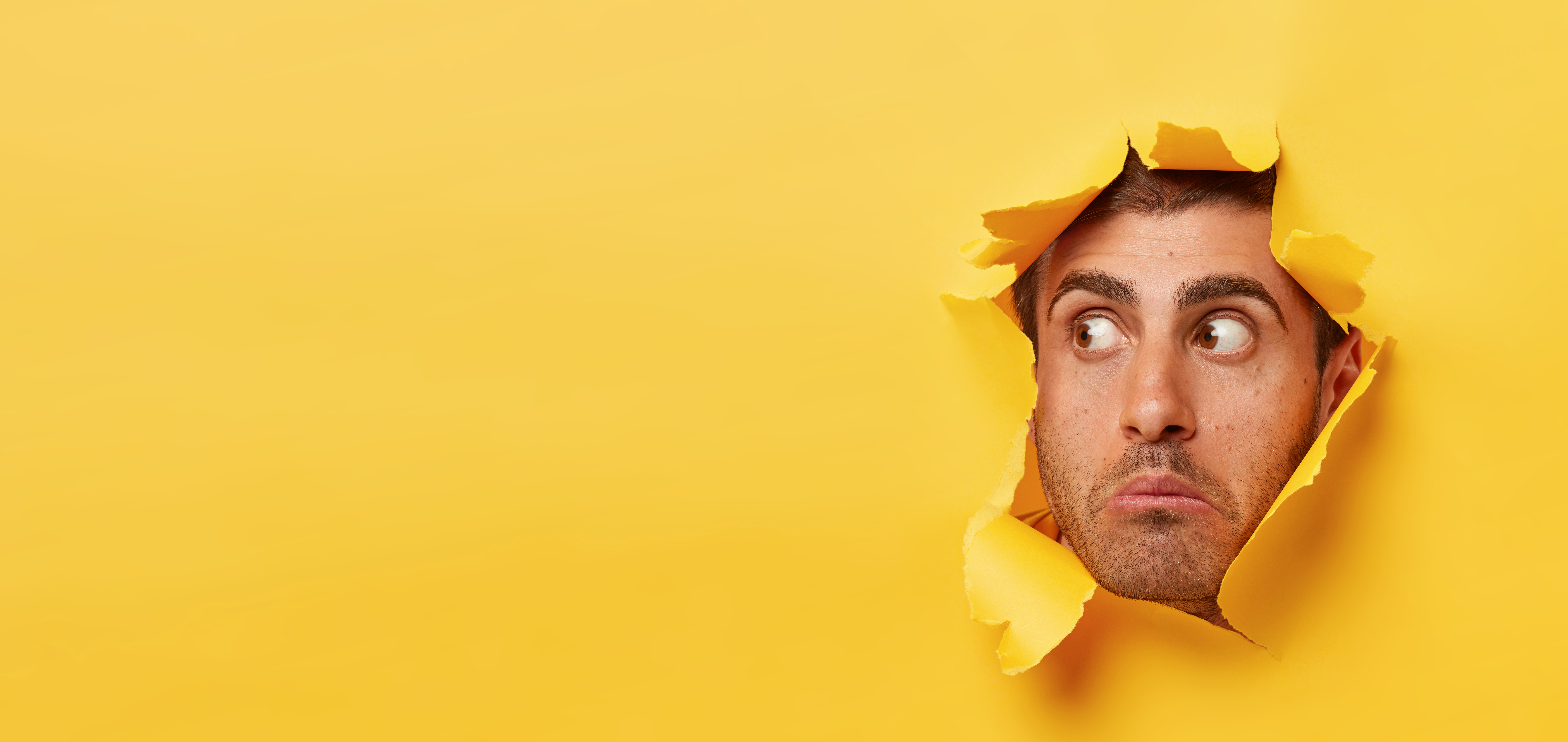 Photo of a man's face pushing through a torn sheet of orange paper. He looks shocked or disappointed.