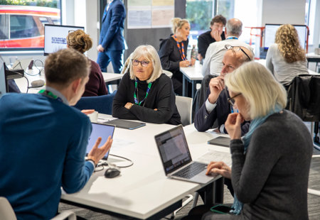 Photo of people sat at a desk talking and listening in an office space