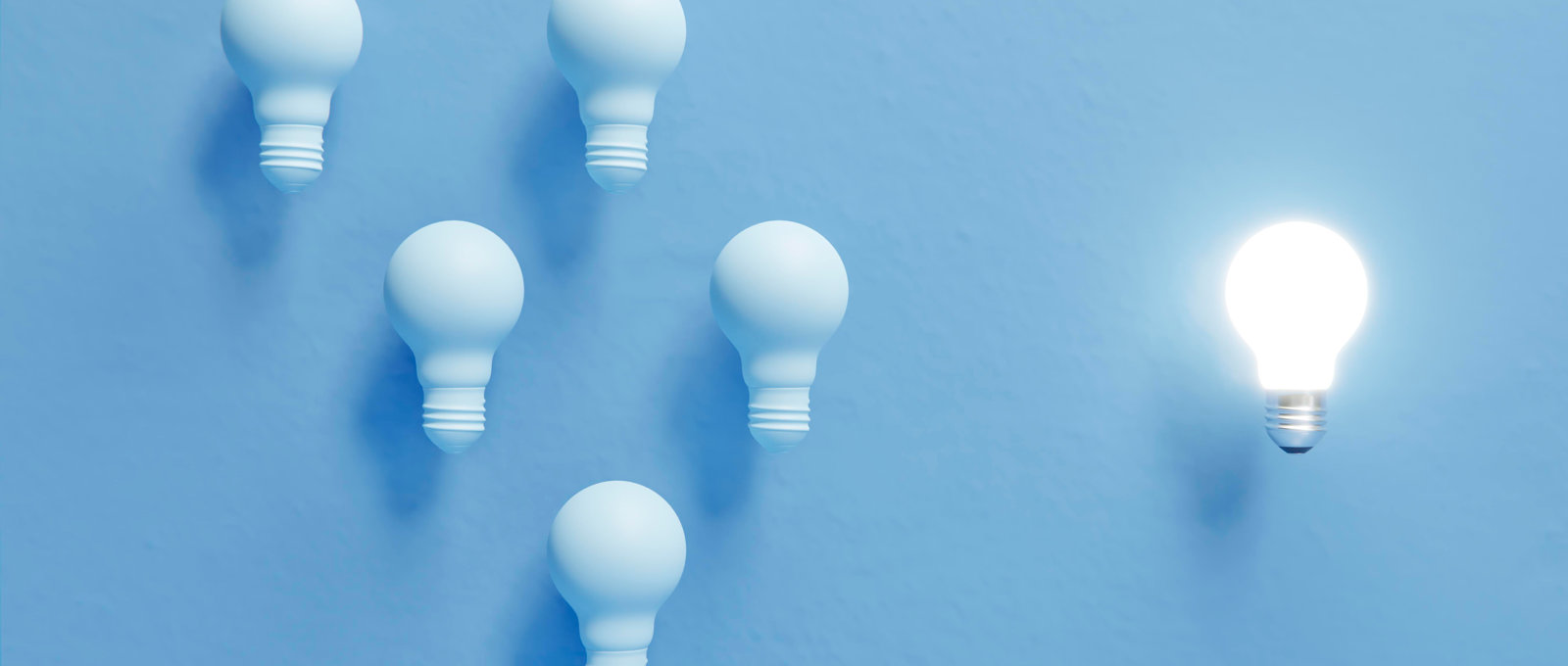 Abstract photo of lightbulbs against a light blue background. Several are dark and grouped together. One is out on its own and lit up.
