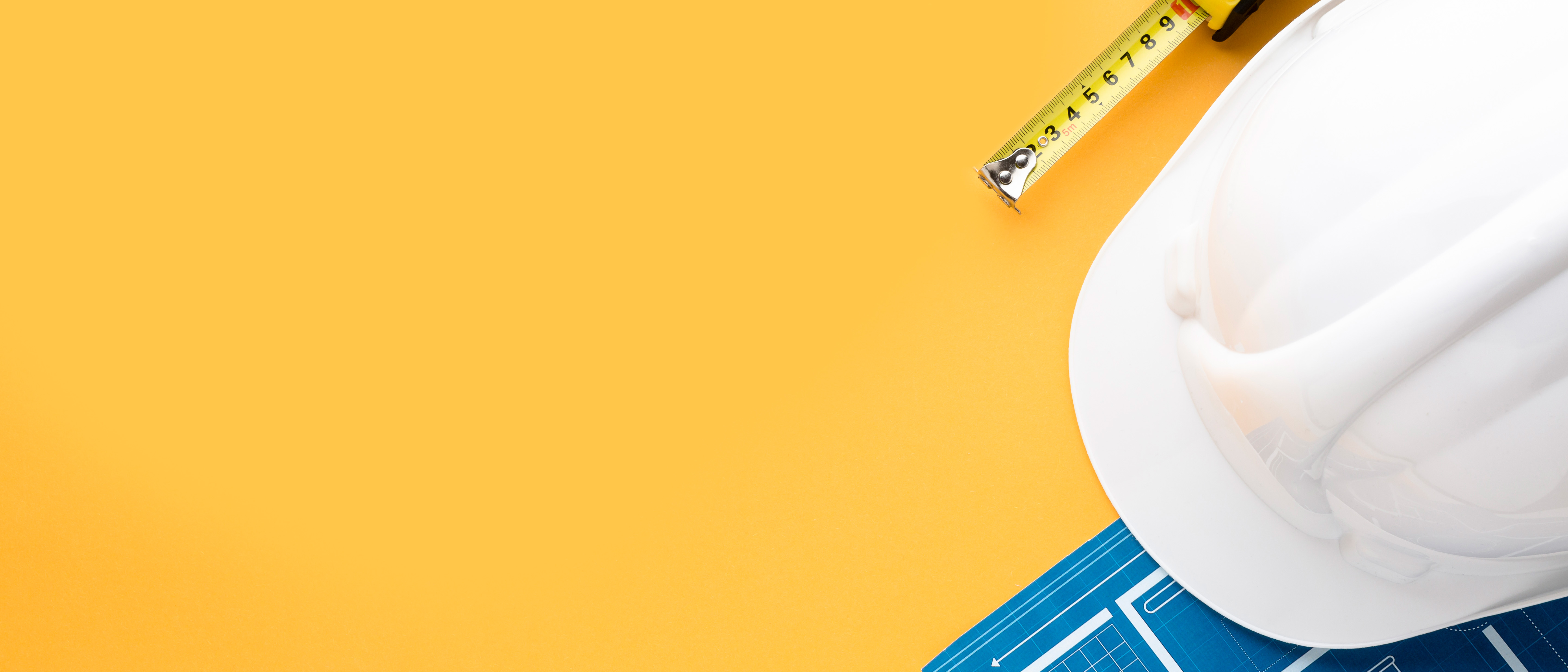 Photo of a white hard-hat, tape measure and some blueprints for a building on a yellow surface