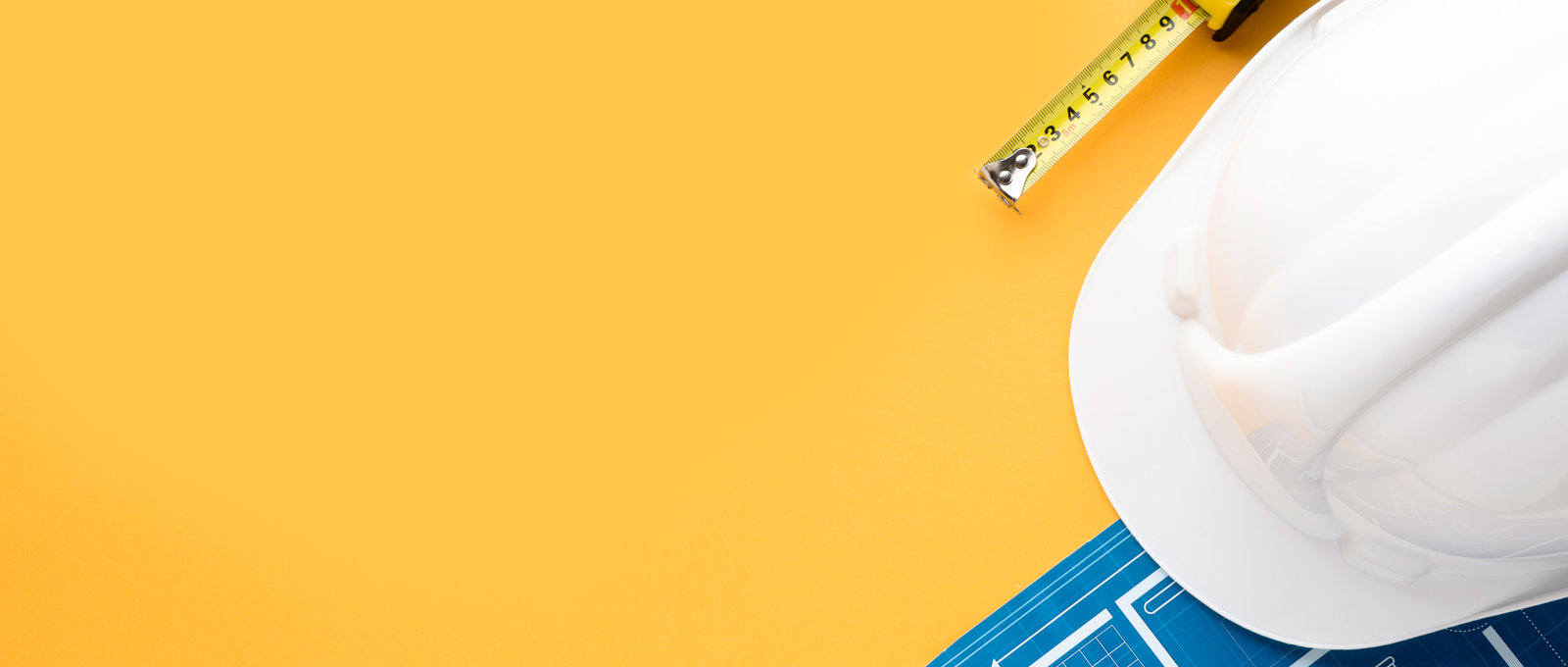 Photo of a white hard-hat, tape measure and some blueprints for a building on a yellow surface