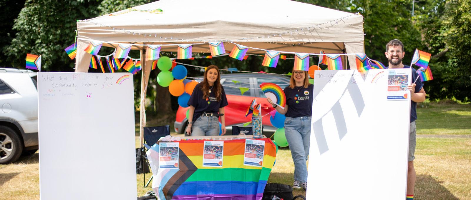 Three members of Futures staff stood around a Pride stall in a Gazebo, flanked with canvases and covered in brightly coloured rainbow flags.