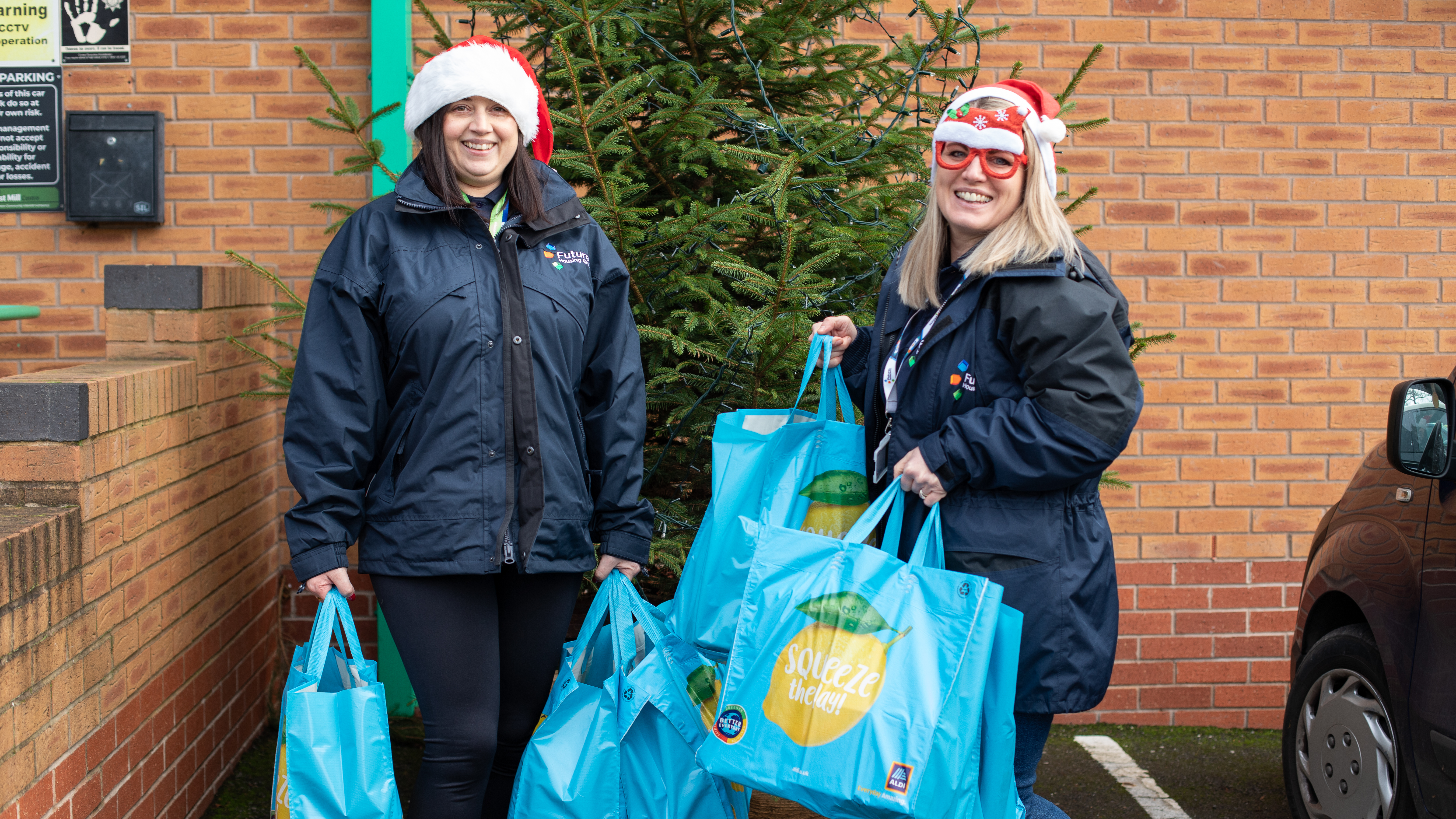 Two female colleagues wearing santa hats, holding shopping bags with food in with a Christmas tree behind them