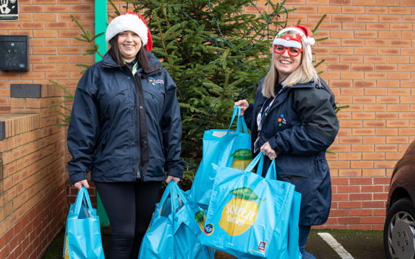 Two female colleagues wearing santa hats, holding shopping bags with food in with a Christmas tree behind them
