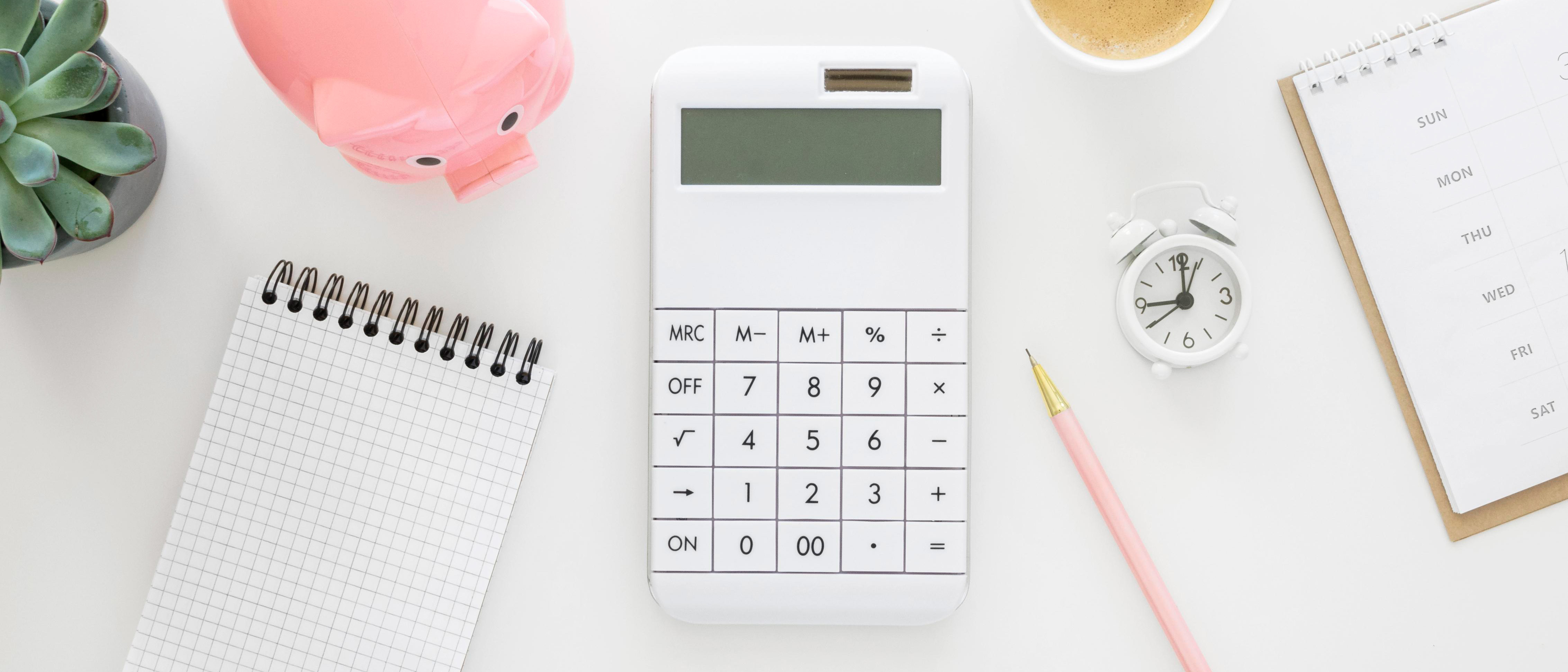 Ariel view of white office desktop with calculator, piggy bank, pen, note pad, diary, calendar and house plant