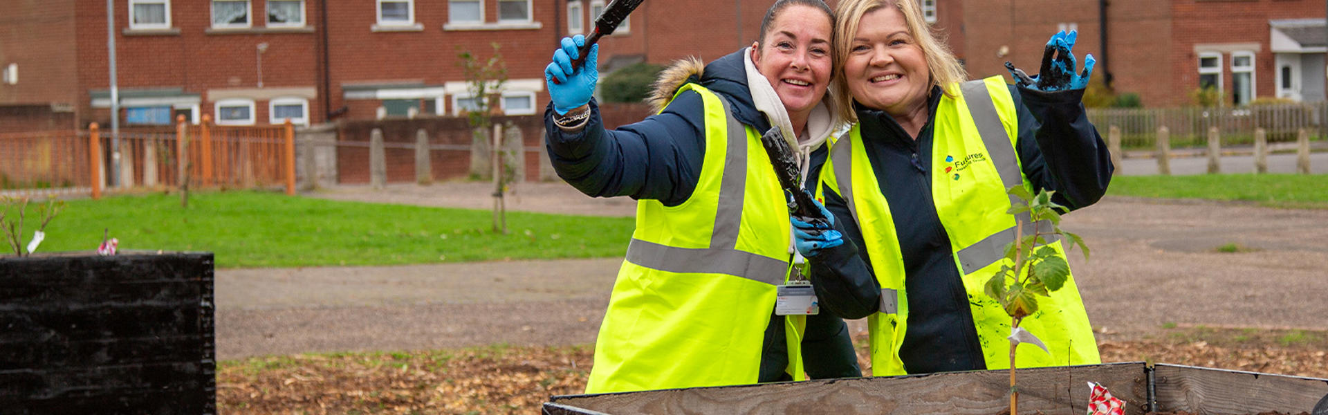Two smiling people in hi-vis jackets posing for a picture holding paintbrushes in front of a planter
