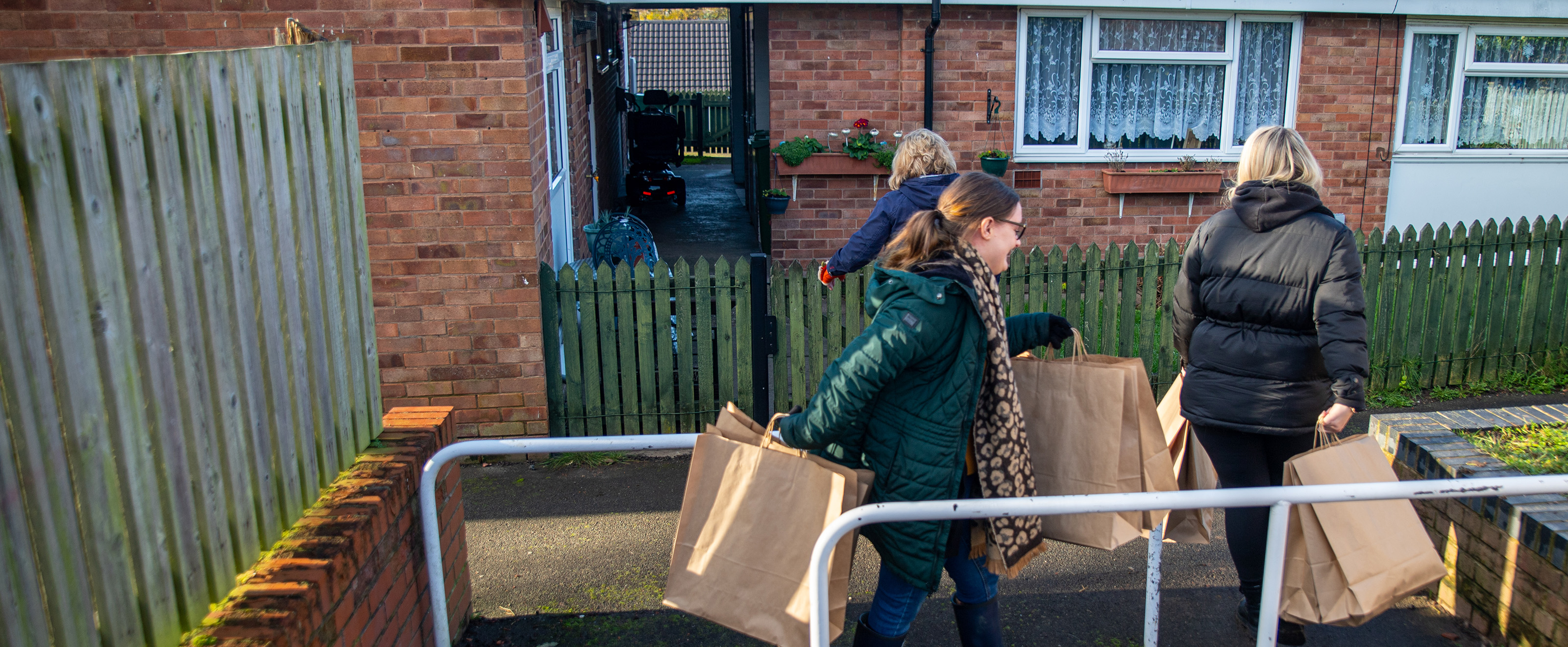 Three women about to enter property through green wooden gate, wearing warm coats and carrying brown paper bags