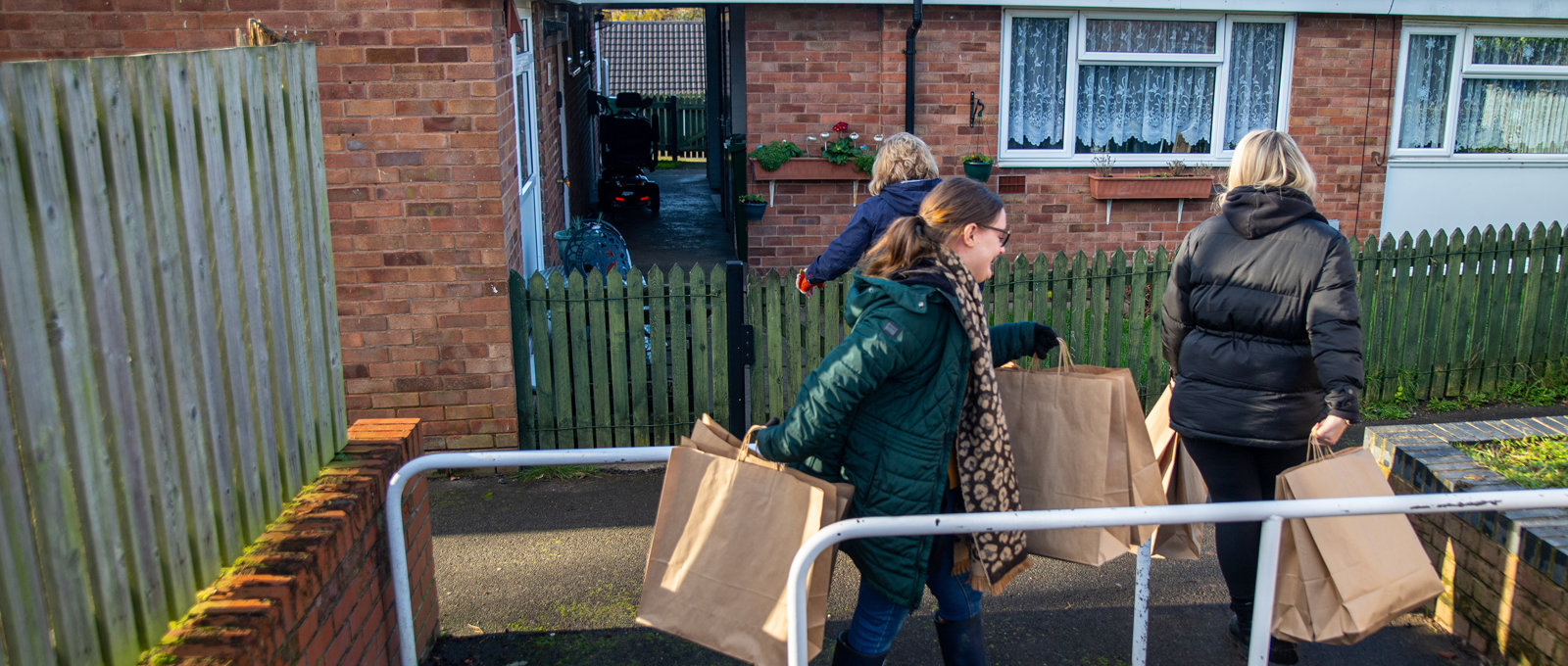 Three women about to enter property through green wooden gate, wearing warm coats and carrying brown paper bags