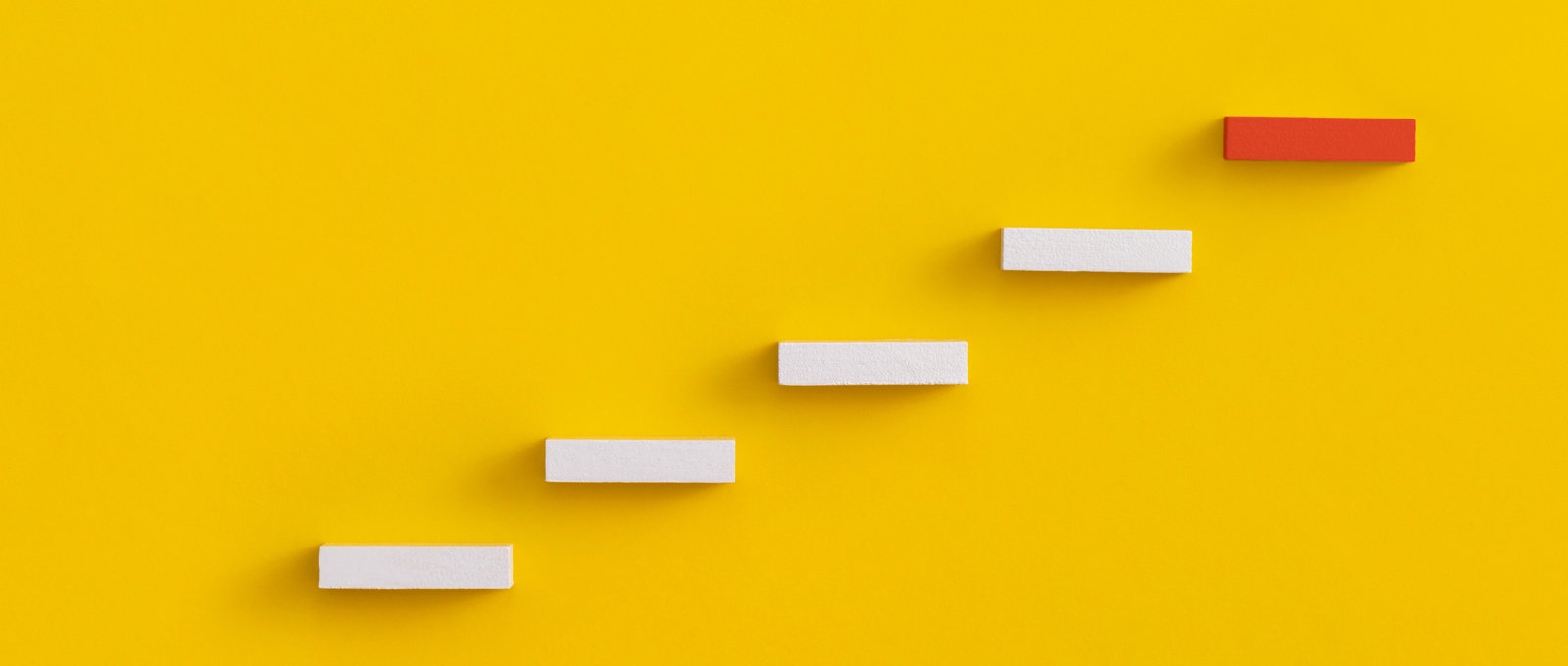 Abstract photo of wooden 'steps' against a yellow background. They go up and right from the bottom left side of the image. The top 'step' is red.