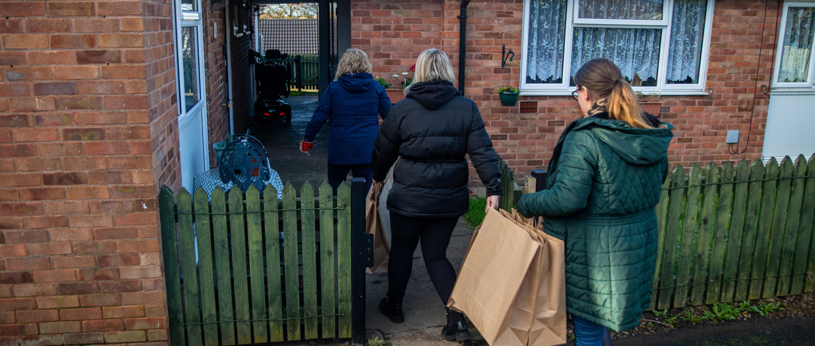 Three women entering property through green wooden gate, wearing warm coats and carrying brown paper bags