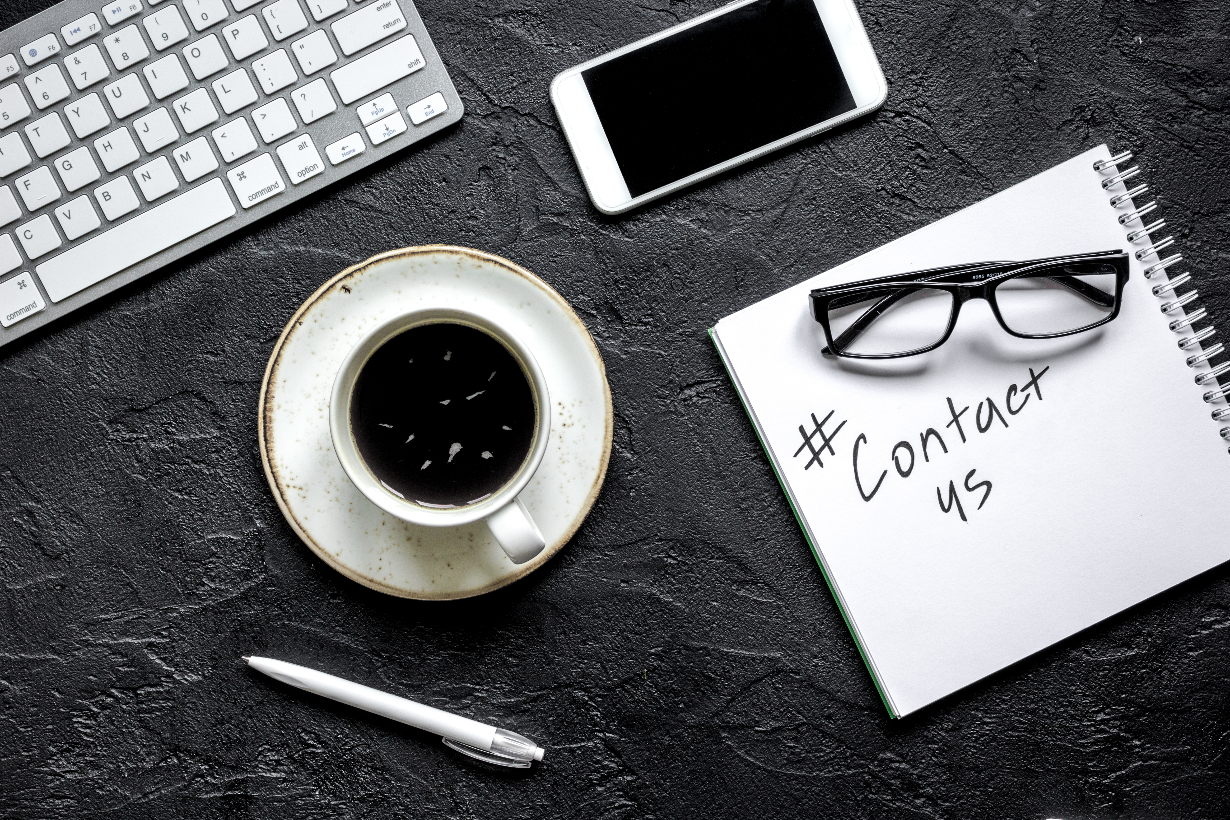 Photograph of a desktop with a keyboard, mobile, cup, pen, spectacles and a notepad on it. On the pad someone has written 'contact us'
