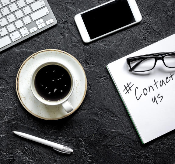 Photograph of a desktop with a keyboard, mobile, cup, pen, spectacles and a notepad on it. On the pad someone has written 'contact us'