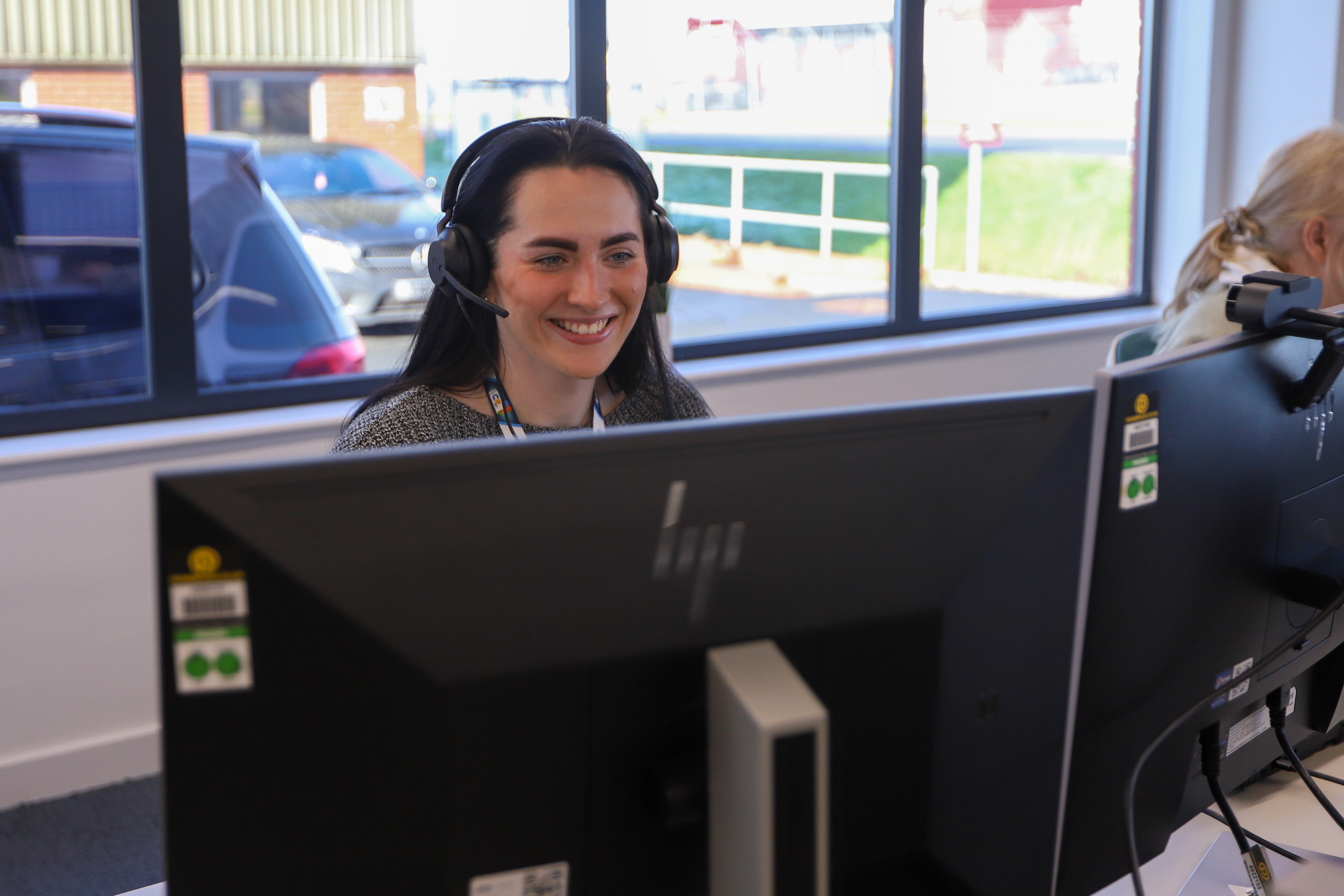 Woman smiling sat at a desk with a computer screen wearing a headset.
