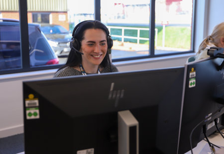 Woman smiling sat at a desk with a computer screen wearing a headset.