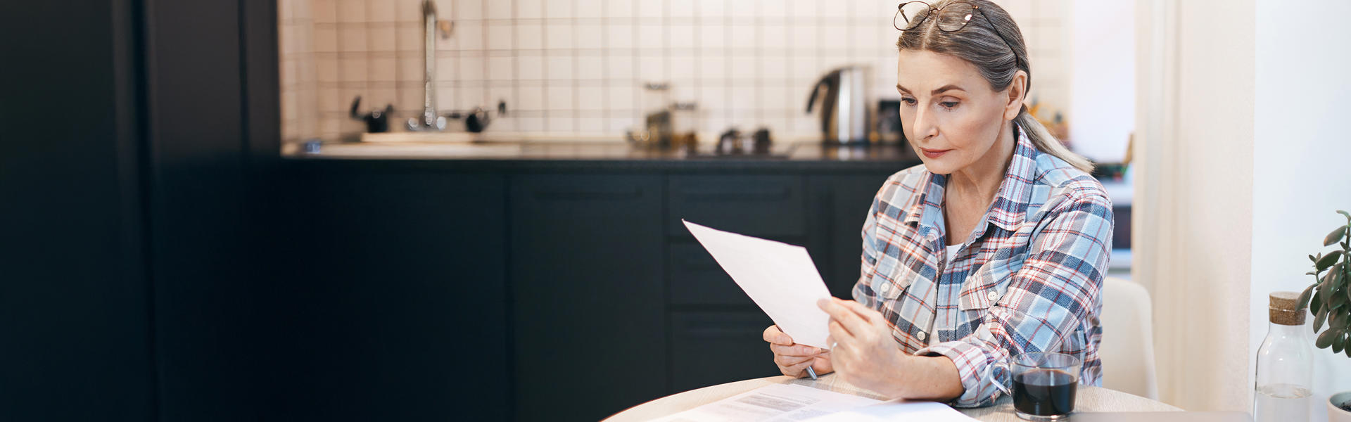 A woman in a plaid shirt sits at a kitchen table, carefully reading a document.