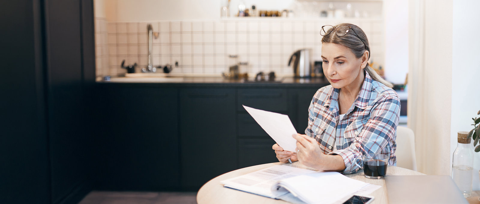A woman in a plaid shirt sits at a kitchen table, carefully reading a document.