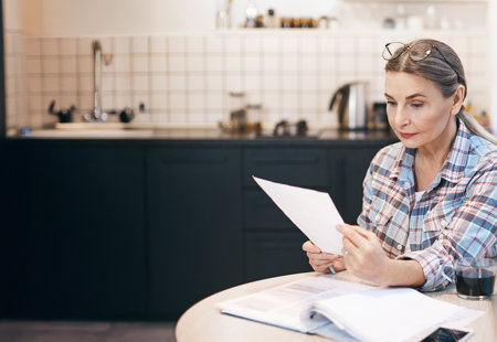 A woman in a plaid shirt sits at a kitchen table, carefully reading a document.