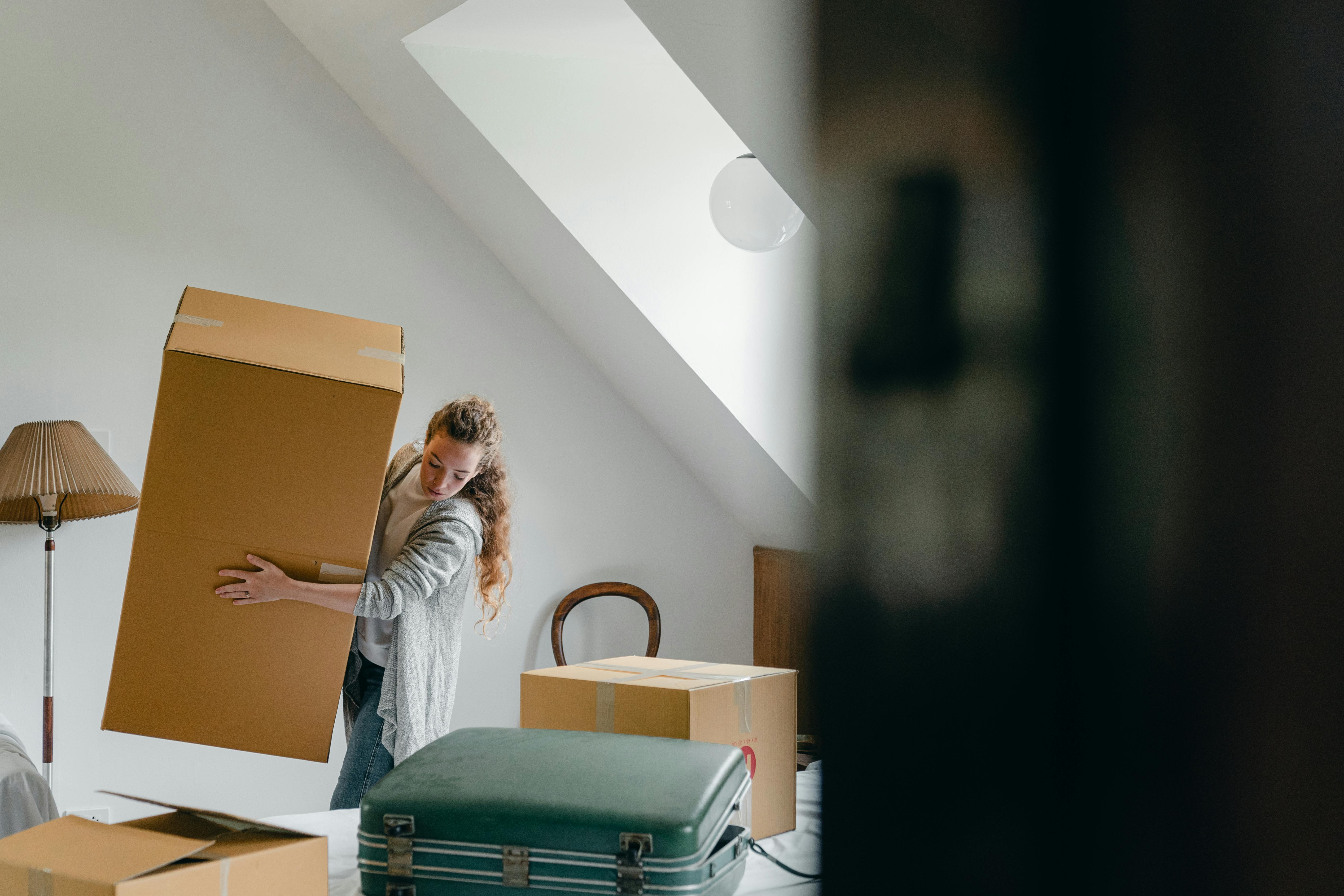 Photo of a woman lifting a large carboard box in what looks like a bedroom. There are other boxes and a suitcase visible