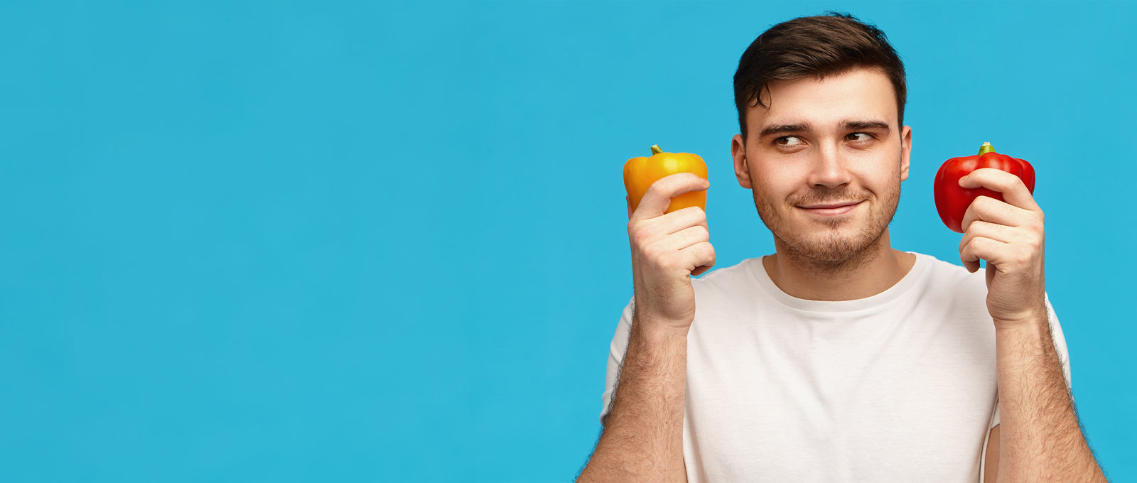 Photo of a man holding a red pepper in one hand and a yellow pepper in the other hand. Standing in front of a blue background