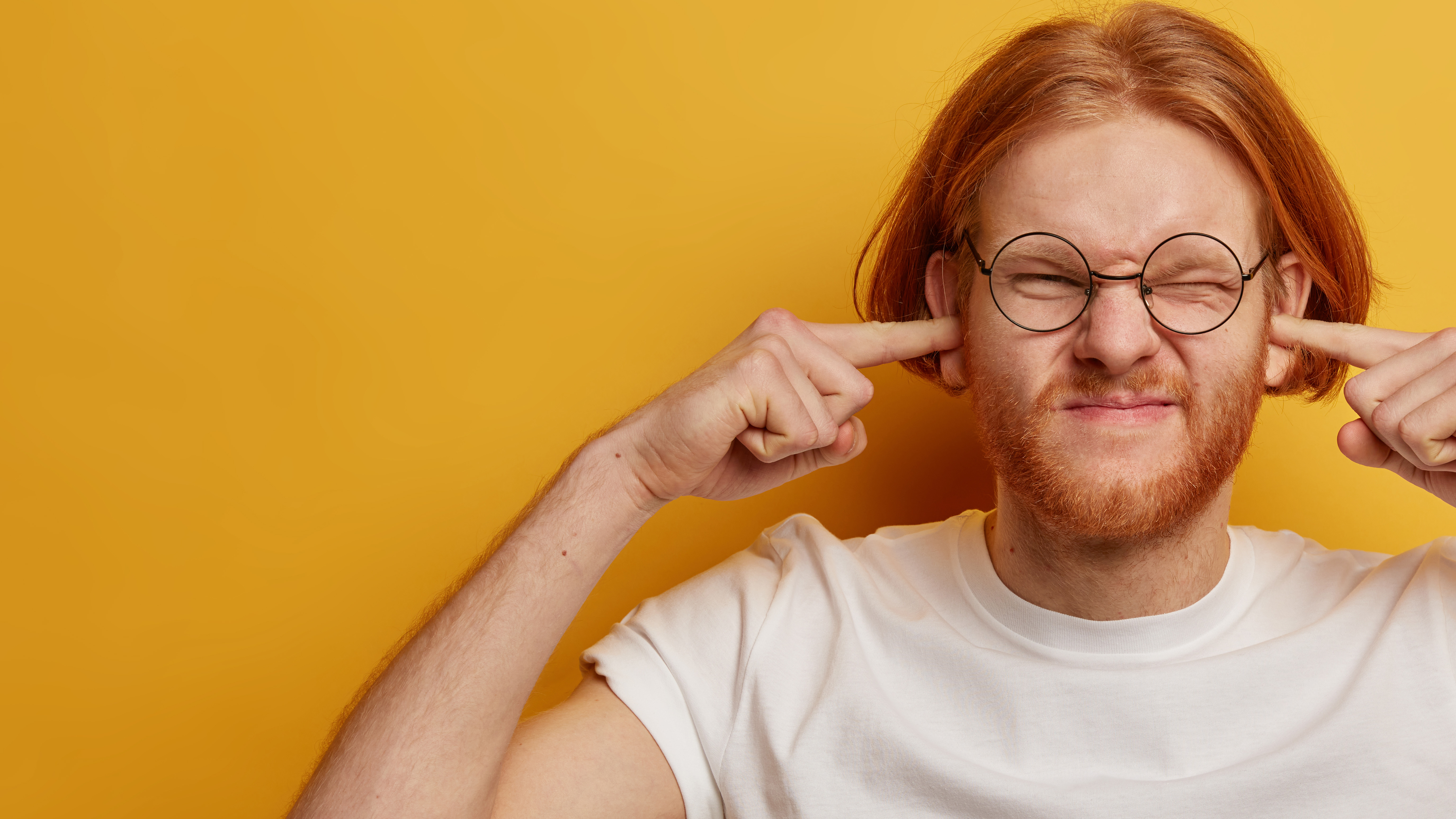 Photo of a young man standing in front of an orange background. He has his fingers in his ears and looks annoyed