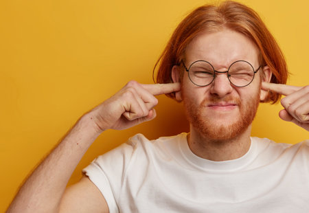 Photo of a young man standing in front of an orange background. He has his fingers in his ears and looks annoyed