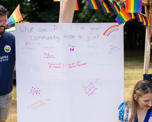 A male member of staff stands next to a canvas reading "What does Pride in your community mean to you", looking down towards a woman wearing blue and with brightly coloured face paint.